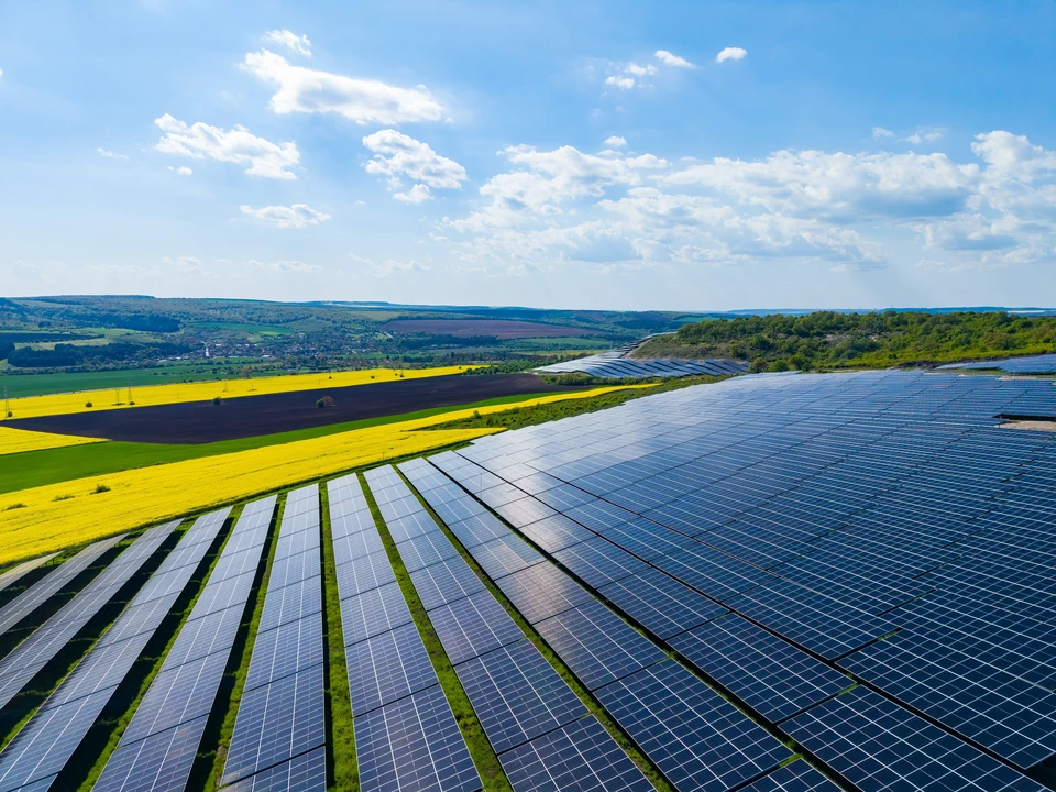 Deux techniciens en combinaisons vertes et casques blancs installent des panneaux photovoltaïques sur une toiture résidentielle en tuiles sous un ciel bleu dégagé, environnement verdoyant visible en arrière-plan