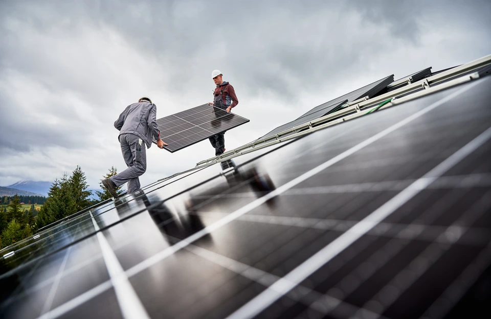 Plan de travail de bureau avec un technicien ou ingénieur étudiant des plans techniques d'installation solaire photovoltaïque sur un écran d'ordinateur ou sur des documents papier étalés sur le bureau. Des schémas techniques, des calculs d'orientation solaire, ou des maquettes d'installation solaire sont visibles. L'environnement doit être professionnel et clair, reflétant la précision technique et l'expertise interne d'un bureau d'études.
