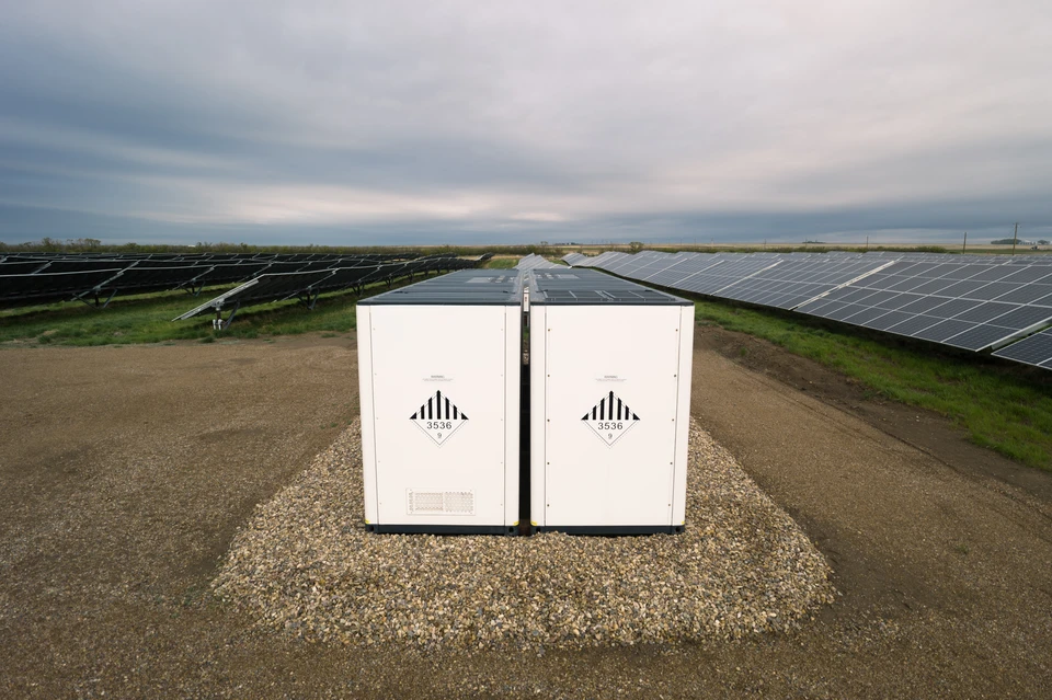 Photographie réaliste d'une batterie de stockage d'énergie solaire montée en intérieur (garage ou cave), connectée à des panneaux solaires visibles via une fenêtre ou une ouverture. La batterie doit être un système moderne de stockage domestique bien identifiable. L'image montre clairement le dispositif de stockage en position d'utilisation, illustrant comment l'énergie solaire générée en journée est conservée pour une utilisation ultérieure. Lumière naturelle mettant en avant le système de batterie et sa fonction de stockage centralisé.